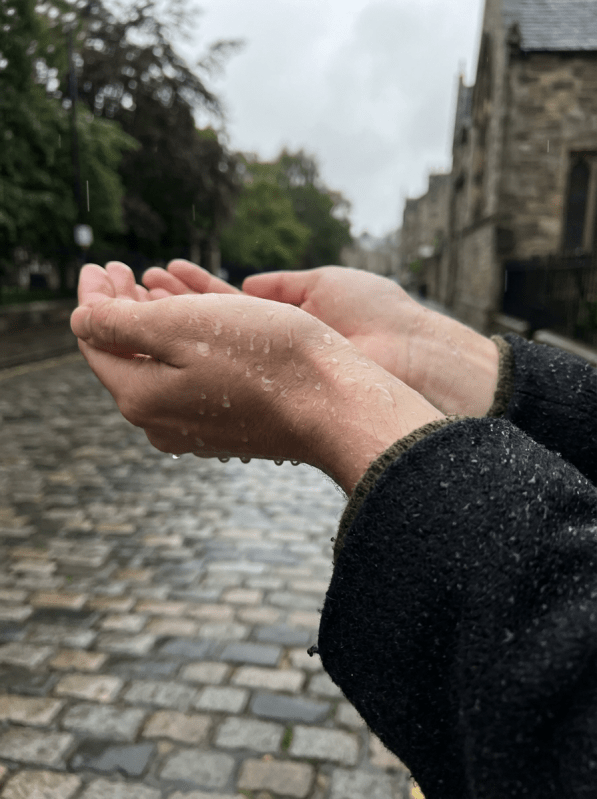 Cupped hands catching rain on a wet cobblestone street with stone buildings in the background.